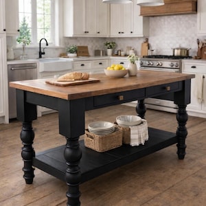 May include: A black and wood kitchen island with a butcher block top, two drawers, and a lower shelf. The island has turned legs and is in a bright, white kitchen with pendant lights and a farmhouse sink. A cutting board with bread and a bowl of lemons are on the island.