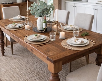 May include: A rectangular wooden dining table with turned legs, set with white plates, woven placemats, linen napkins, gold-toned cutlery, clear glasses, pillar candles in glass holders, and a central wire basket holding a white vase with greenery.