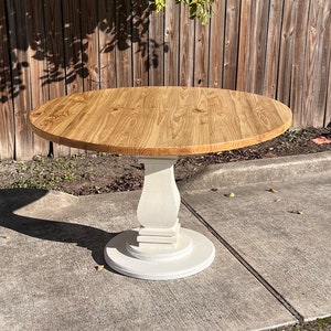 May include: A round dining table featuring a natural light brown wood grain top with visible circular patterns. The table is supported by a central pedestal base painted in a solid matte white finish, consisting of a square column and a round base.