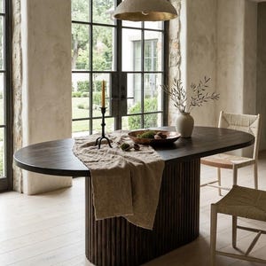 May include: An oval dark wood dining table with a fluted pedestal base, draped with a beige linen runner. The table holds a black metal candle holder with a yellow taper candle, a copper bowl with greenery, and a white vase with olive branches.