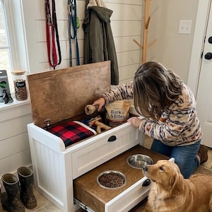 May include: A white dog food storage bench with a pull-out drawer containing food and stainless steel bowls. The bench has a wooden top and a red and black plaid blanket inside. A golden retriever dog is lying on a rug.