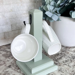 May include: A light green wooden mug holder with two white ceramic mugs hanging on hooks. The holder has a square base and a vertical post with two hooks.