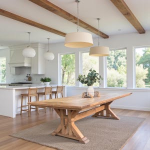 May include: A large, light-colored wooden dining table with a trestle base. The table is set on a neutral-toned rug in a bright, open kitchen. Overhead, two pendant lights hang above the table, and the room features large windows.