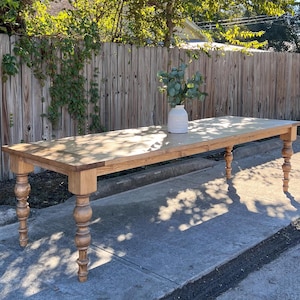 May include: A long, wooden farmhouse table with a natural finish and turned legs. The table is set on a paved driveway with a wooden fence in the background.