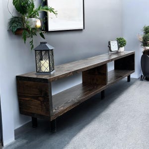 May include: A dark brown wooden media console with three open shelves and black metal legs. The console is 60 inches long and 16 inches deep.