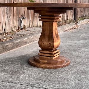May include: A round wooden table with a dark brown stained finish. The table features a central pedestal base with a curved, urn-shaped support column, accented by square tiered blocks at the top and bottom, resting on a circular flat base.