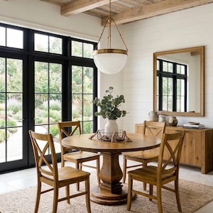 May include: A round wooden dining table with a pedestal base, surrounded by four wooden chairs with x-back designs. A white vase with green foliage sits in a wire basket on the table, beneath a large brass and glass pendant light fixture.
