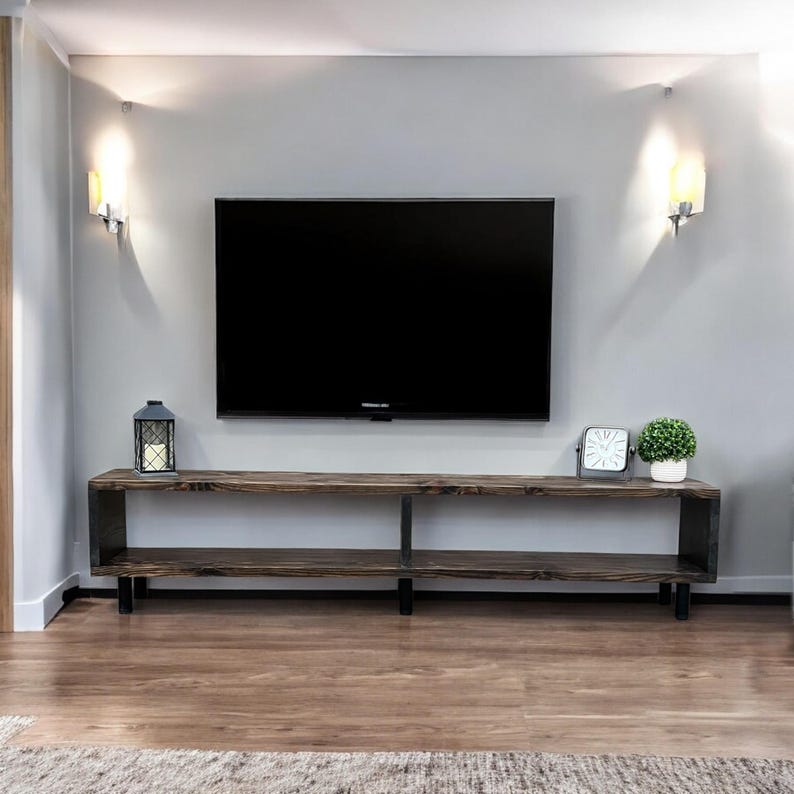 May include: A dark wood TV stand with two shelves and black metal legs. The stand is in front of a gray wall with two wall sconces on either side of a flat screen TV.