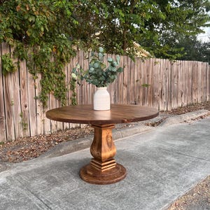 May include: A round wooden dining table with a dark brown stained finish and a central pedestal base. The table features a smooth circular top and a sturdy, carved column support resting on a round base. A white vase with green foliage sits on top.