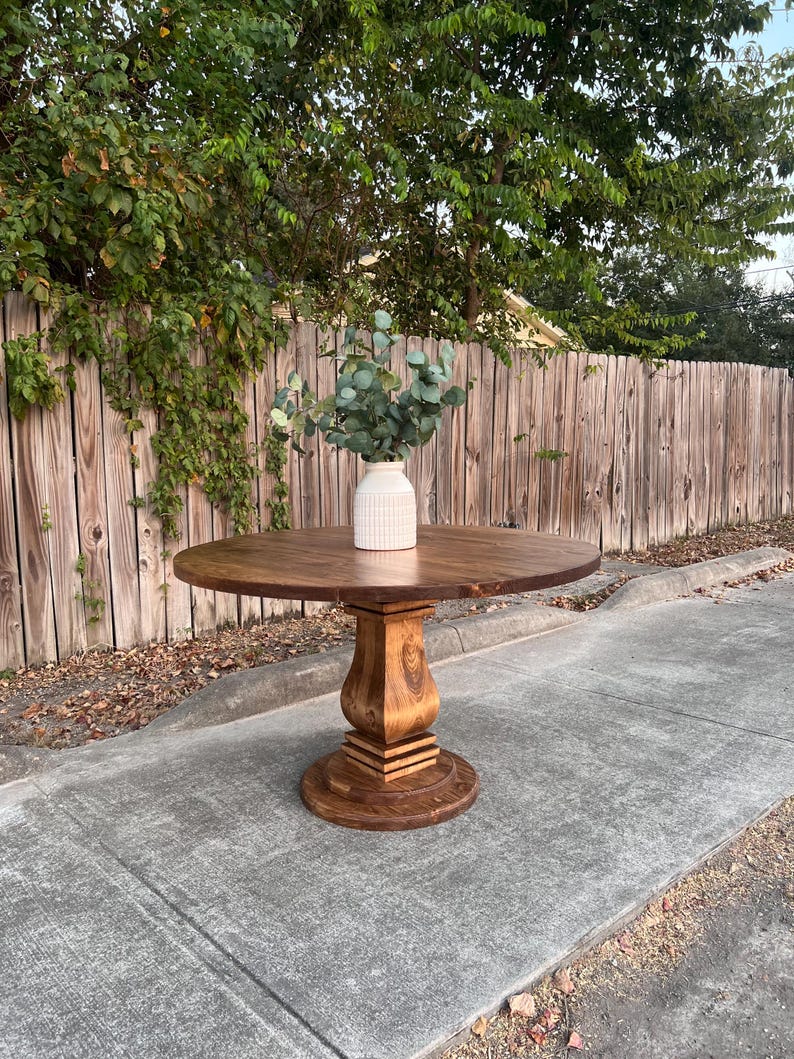 May include: A round wooden dining table with a dark brown stained finish and a central pedestal base. The table features a smooth circular top and a sturdy, carved column support resting on a round base. A white vase with green foliage sits on top.