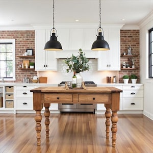 May include: A rustic wooden kitchen island with two drawers and turned legs. The island is topped with a vase of greenery and a bottle of wine in a wire basket. Two black pendant lights hang above the island, and the kitchen features white cabinets and brick accents.