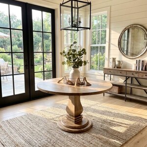 May include: A round wooden pedestal table sits on a textured jute rug. On the table is a white vase with green foliage inside a wire basket with decorative tassels. Above hangs a black metal lantern pendant light in a room with white walls.