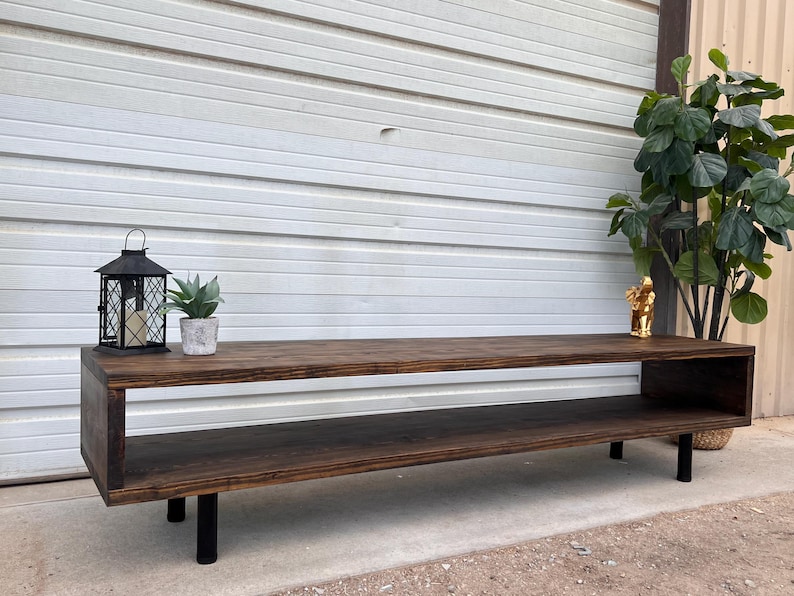 May include: A dark brown wooden coffee table with a lower shelf and black metal legs. The table is sitting in front of a white wall and has a lantern and a potted plant on top.