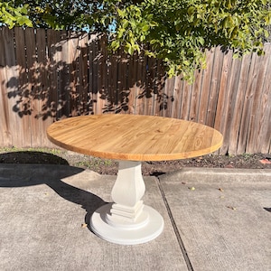 May include: A round dining table featuring a natural light brown wood grain top with a smooth finish. The table is supported by a single, thick, off-white pedestal base that flares out at the bottom into a circular foundation for stability.