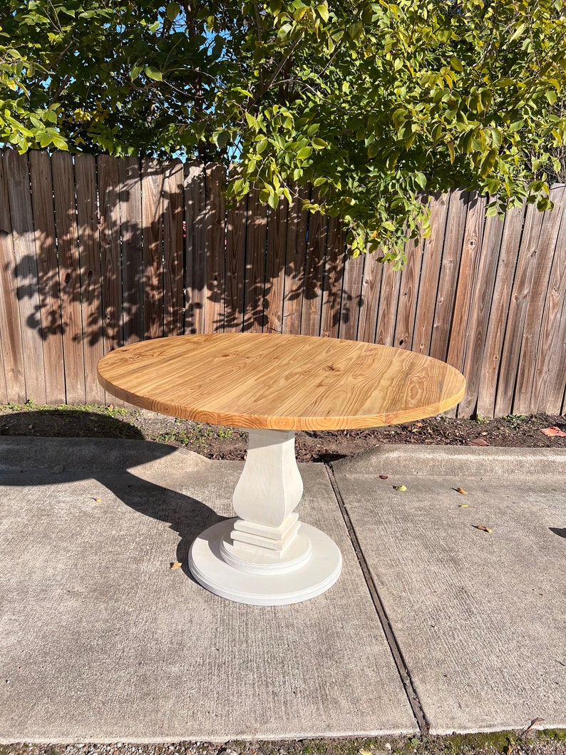 May include: A round dining table featuring a natural light brown wood grain top with a smooth finish. The table is supported by a single, thick, off-white pedestal base that flares out at the bottom into a circular foundation for stability.