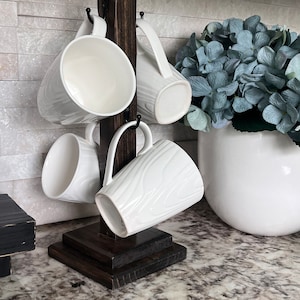 May include: A dark wood mug rack with four white ceramic mugs hanging from hooks. The rack has a square base and is sitting on a granite countertop.
