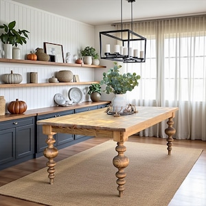 May include: A light-colored wooden dining table with turned legs is the focal point of a well-lit room. The table is rectangular and sits on a beige rug. Shelves with decorative items and a black chandelier complete the scene.