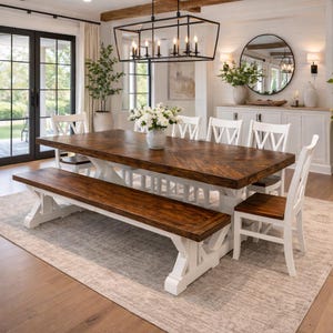 May include: A large, rectangular dining table with a dark brown, chevron-patterned top and white base. A matching bench sits on one side, with white chairs around the table. The room has a neutral color palette with a rug and a chandelier.