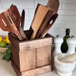May include: A rustic wooden utensil holder filled with various wooden kitchen tools. The holder is made of stained wood and sits on a countertop with other kitchen items, including salt and pepper shakers, a mortar and pestle, and a bottle of olive oil.