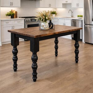 May include: A rectangular kitchen island with a dark wood top and black legs. The legs are turned and have a decorative, rounded design. The island is in a bright kitchen with white cabinets and stainless steel appliances. The island is approximately 48 inches long.