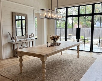 May include: A large rectangular wooden dining table with turned legs sits on a textured jute rug. Above the table hangs a rectangular black metal chandelier with multiple light bulbs. The room features white shiplap walls and large black-framed windows.