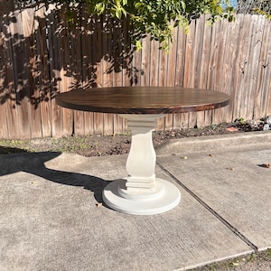 May include: A round dining table featuring a dark brown stained circular top supported by a central pedestal base. The pedestal and the circular floor plate are finished in a matte off-white color, providing a contrast to the dark wood surface.