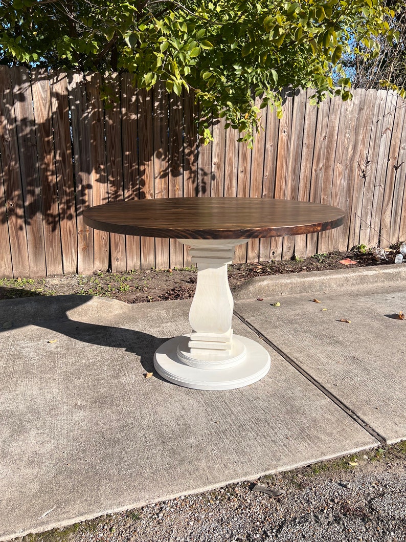 May include: A round dining table featuring a dark brown stained circular top supported by a central pedestal base. The pedestal and the circular floor plate are finished in a matte off-white color, providing a contrast to the dark wood surface.