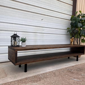 May include: A dark brown wooden media console with a shelf and black metal legs. The console is sitting on a gravel surface in front of a white garage door.