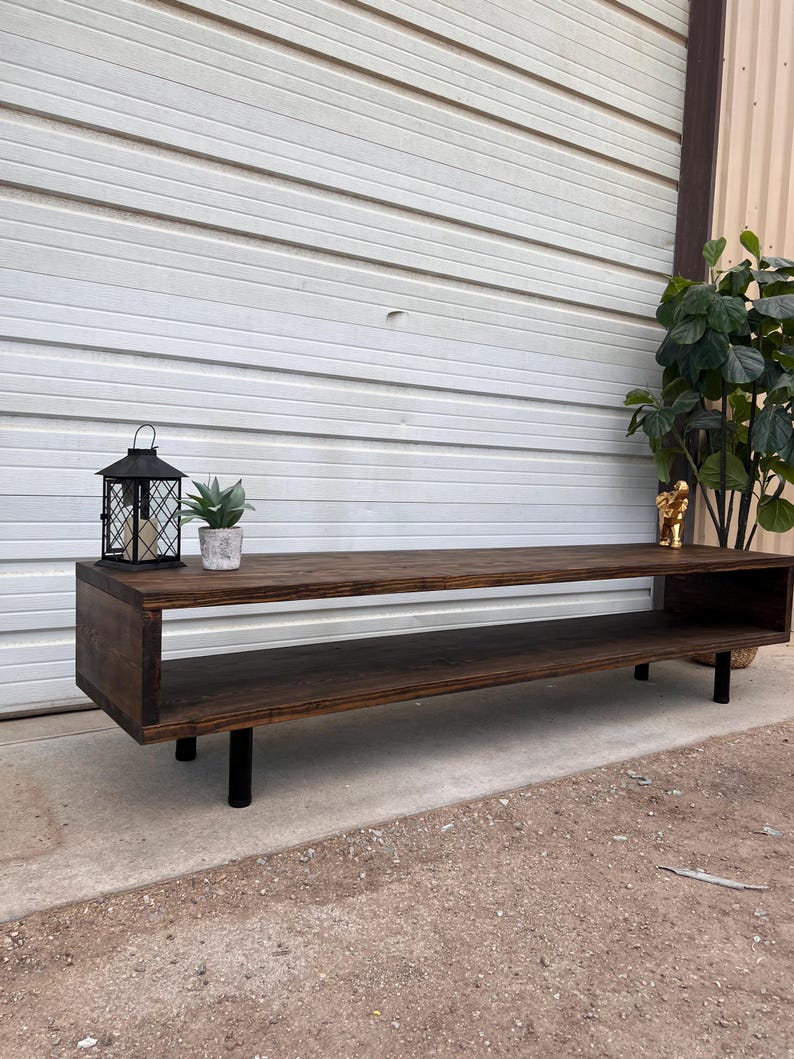 May include: A dark brown wooden media console with a shelf and black metal legs. The console is sitting on a gravel surface in front of a white garage door.