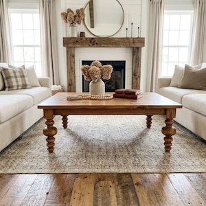 May include: A rectangular wooden coffee table with four turned legs, positioned between two beige sofas. The table surface holds a ceramic vase with dried flowers, a wooden bead garland, and two stacked brown books, all set on a patterned rug.