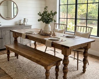 May include: A rectangular wooden dining table with turned legs, accompanied by a matching wooden bench and two spindle-back chairs. The table is set with plates, napkins, and a central white vase containing green foliage on a woven rug.