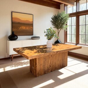 May include: A rectangular wooden dining table with a thick slab top and a solid block base. The table features a natural wood grain finish and is positioned in a bright room with a white sideboard, a black vase, and a white vase with greenery.