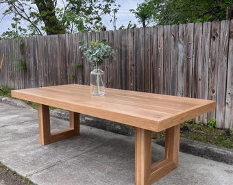 May include: A long, rectangular wooden dining table with a natural wood finish and simple square legs. The table is made of solid wood and has a smooth, polished surface. The table is sitting on a concrete sidewalk.