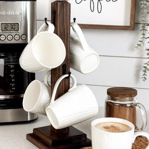 May include: A dark wood mug tree holds six white ceramic mugs with a textured design. A coffee maker and a sign that says "coffee" are in the background. A mug of coffee sits on a woven coaster.