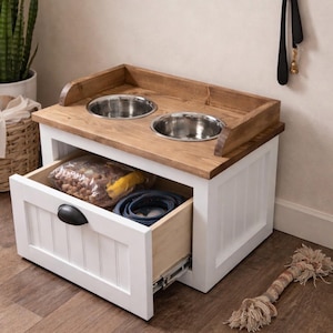 May include: A white and wood pet feeding station with two stainless steel bowls. The unit has a drawer that is open, revealing pet supplies. A dog toy is on the floor.