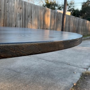 May include: A circular wooden table top with a dark brown stained finish. The surface displays a visible wood grain pattern, and the rounded edge shows the texture of the timber. The piece is positioned horizontally, highlighting the smooth, flat top.