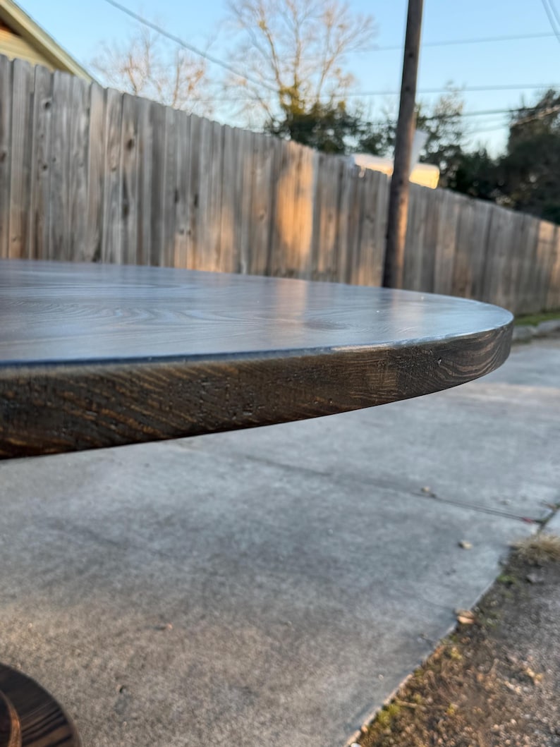 May include: A circular wooden table top with a dark brown stained finish. The surface displays a visible wood grain pattern, and the rounded edge shows the texture of the timber. The piece is positioned horizontally, highlighting the smooth, flat top.