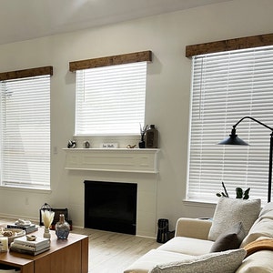 May include: A living room with a white fireplace, a couch, and three windows with white blinds and wooden beams above them. The windows are covered with white blinds. The couch is white with a brown throw pillow and a tan throw blanket. The fireplace has a black firebox. The room is painted white.