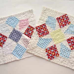 May include: Two square quilted pot holders. Each has a white background with colorful patchwork squares. The squares are blue gingham, red with white dots, pink, yellow, and blue polka dots. The pot holders are made of fabric.