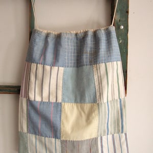 May include: A patchwork bag with a drawstring closure. The bag is made of various fabric squares in shades of blue, white, and gray, with striped and checkered patterns. The bag hangs from a wooden structure with a light-colored cord.