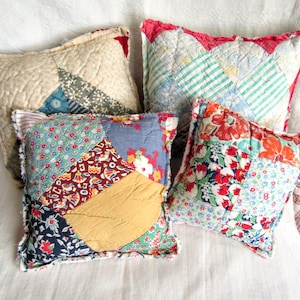 May include: Four square throw pillows made from various quilted fabric patches. The pillows feature diverse patterns including floral prints, stripes, and solid colors in shades of red, blue, yellow, and white, with visible stitching throughout the surfaces.