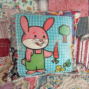 May include: A square pillow with a cartoon bunny holding a carrot. The bunny is pink with green overalls, set against a blue gingham background. The pillow has a patchwork border with floral and patterned fabrics. A tree and flowers are also on the pillow.