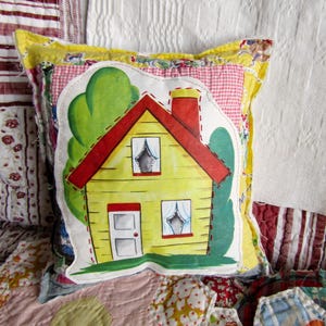 May include: A decorative pillow featuring a yellow house with a red roof, chimney, and green trees. The house design is stitched onto a patchwork pillow with a yellow and red gingham border. The pillow is on a patchwork quilt.