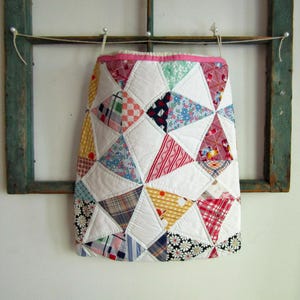 May include: A quilted fabric pouch featuring a patchwork design of various patterned triangles in red, blue, yellow, and black, interspersed with solid white triangles. The pouch has a bright pink top trim and is suspended by a thin white string.