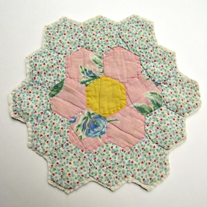 May include: A quilted fabric mat constructed from hexagonal patches. The center is a solid yellow hexagon surrounded by pink floral hexagons, with an outer border of small blue and red floral patterns on a white background with white stitching.