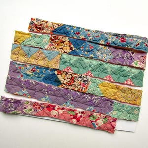 May include: A collection of five long, narrow strips of quilted fabric arranged in parallel. Each strip features a patchwork design composed of various floral and geometric patterns in shades of blue, yellow, green, purple, and pink.