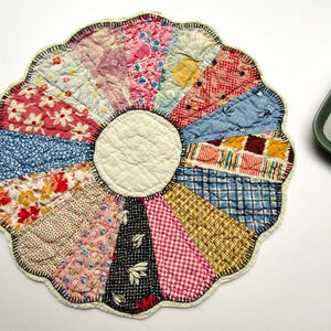 May include: A circular quilted fabric mat featuring a central cream-colored circle surrounded by sixteen wedge-shaped panels in various floral, plaid, and checkered patterns in red, blue, yellow, black, and pink, finished with a scalloped edge.