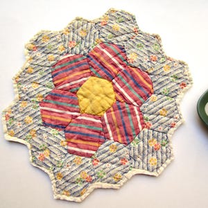 May include: A quilted fabric potholder shaped like a flower with a central yellow hexagon surrounded by six pink and white striped hexagons, and an outer layer of blue and white patterned hexagons with small floral accents and cream trim.