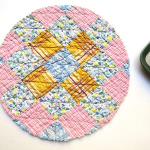 May include: A round, quilted hot pad with a patchwork design. The pad features squares of pink gingham, floral patterns in blue and yellow, and gold and blue squares. The overall design is circular and the pad is likely used for kitchenware.
