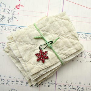 May include: A stack of off-white, textured, rectangular fabric pieces tied together with a thin light green string. A dark green safety pin is attached to the string, holding a small, bright red snowflake-shaped metal charm.